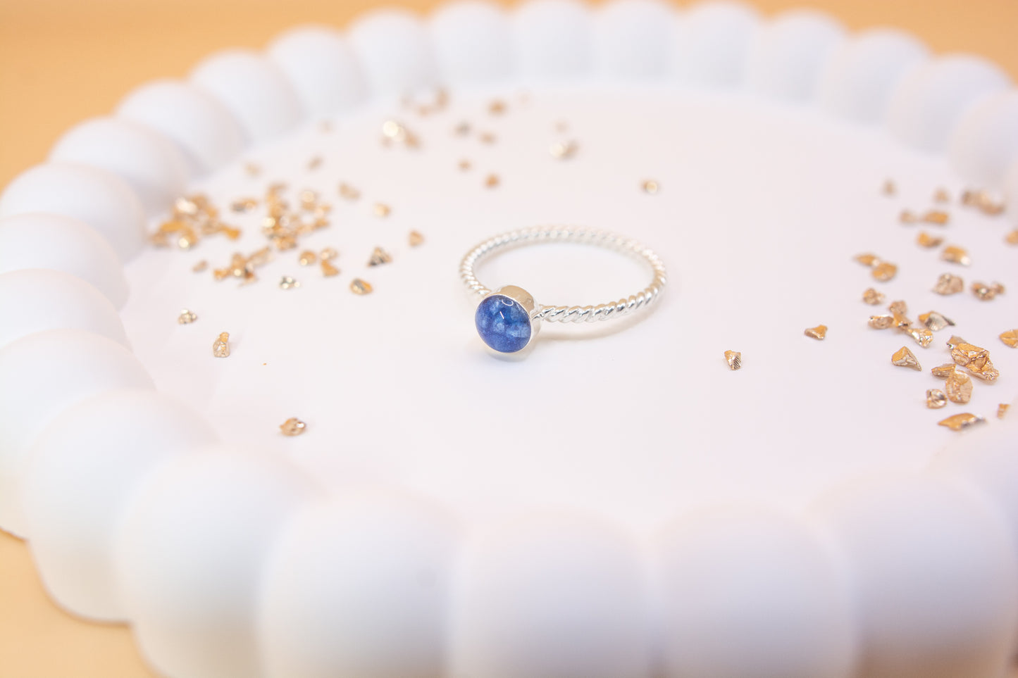 A 45 degree angled view of a twist design 925 sterling silver remembrance ring, displayed on a white jewelry plate with gold fragments either side of it. The ring is cobalt blue in colour and contains cremation ashes