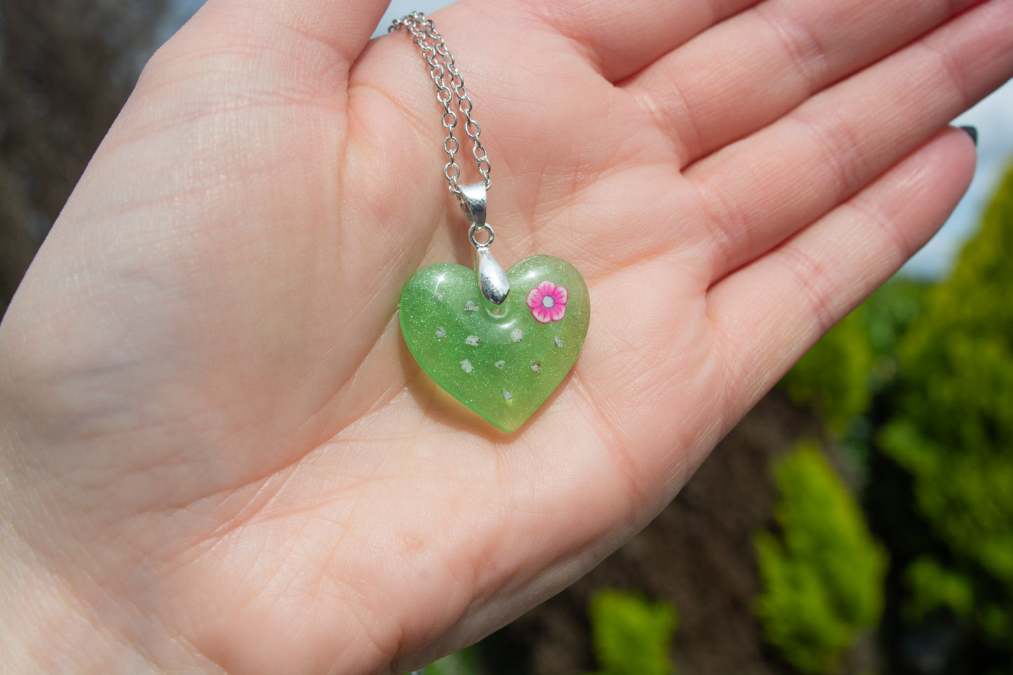 A hand holding a green heart keepsake memorial pendant outside, containing a flower decoration and cremation ashes, set on a 925 sterling silver chain