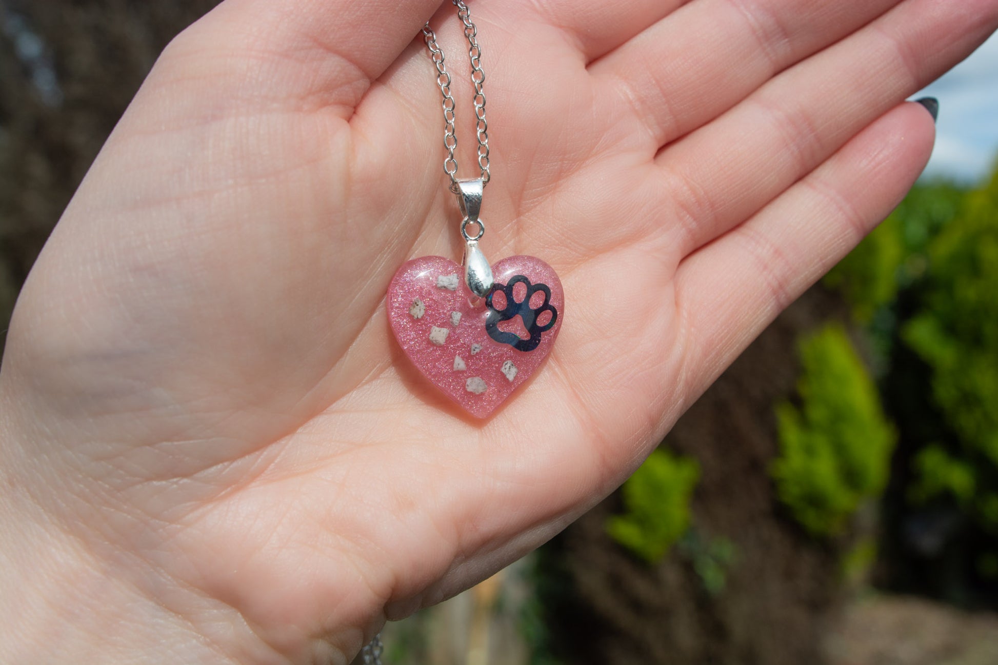 A hand presenting a heart shaped keepsake memorial pendant outside, set on a 925 sterling silver chain, containing a paw print decal and pet cremation ashes