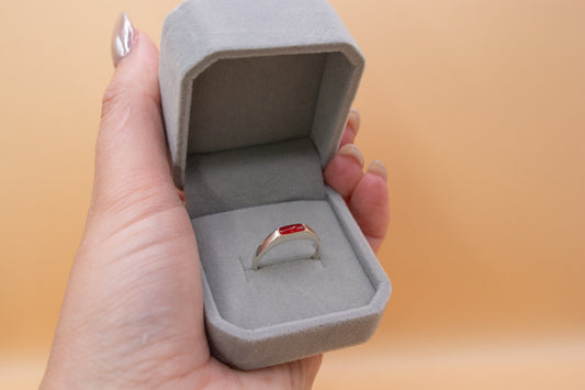 A hand holding a grey plush ring box, inside is a sterling 925 silver ring with a rectangle shaped bezel, containing cherry red resin and cremation ashes