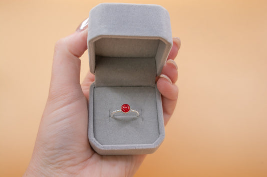 A hand holding a grey plush ring box, inside is a sterling 925 silver twist ring with a round bezel containing cherry red resin and cremation ashes