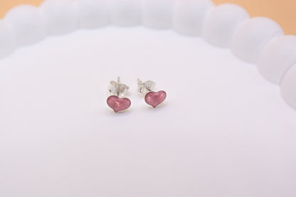 A close up shot of a pair of 925 sterling silver studs, in a heart shape and metallic light pink in colour, containing cremation ashes