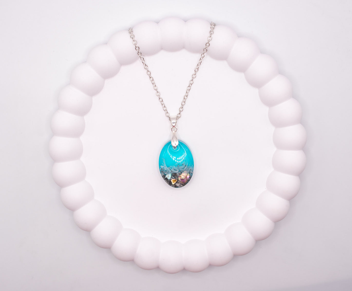 An overhead view of an Oval shaped seashore memorial keepsake pendant, containing cremation ashes and shell pieces, presented on a sterling 925 silver chain