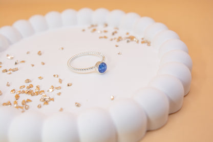 A side view of a twist design sterling silver memorial ring, displayed on a white jewelry plate with gold fragments either side of it. The ring is cobalt blue in colour and contains cremation ashes