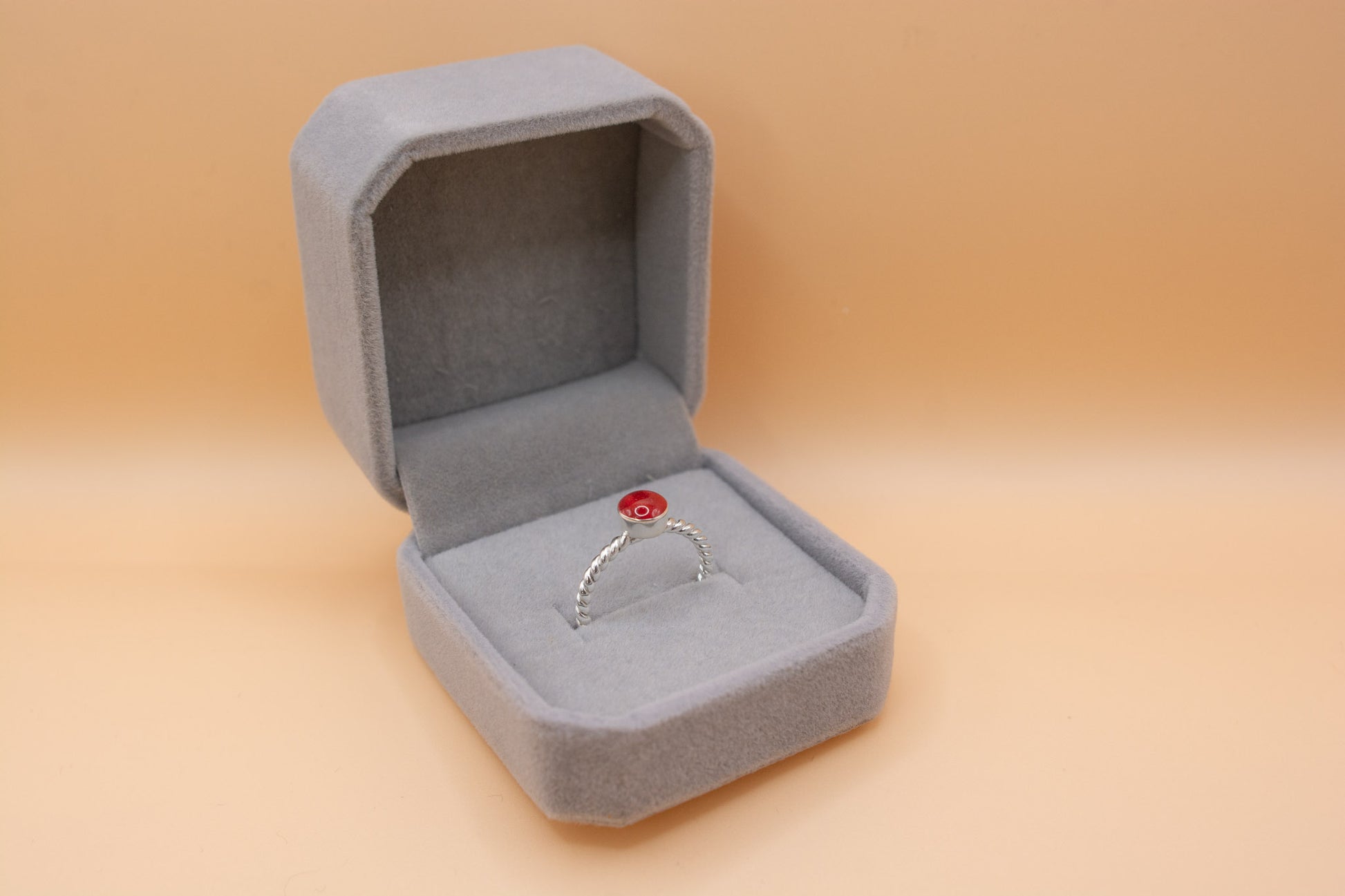 An angled view of a plush grey jewelry box, containing a 925 silver twisted band ring, styled with cherry red resin and containing pet cremation ashes