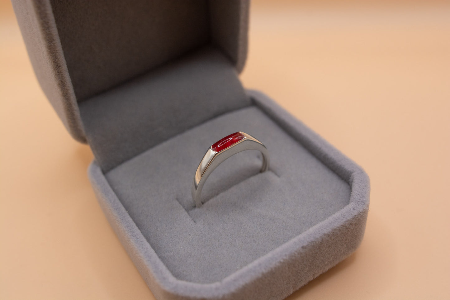 A close-up angled view of a solid 925 sterling silver ring with a rectangle bezel, inside the bezel is red resin and pet cremation ashes. The ring is presented in a grey plush jewelry box.