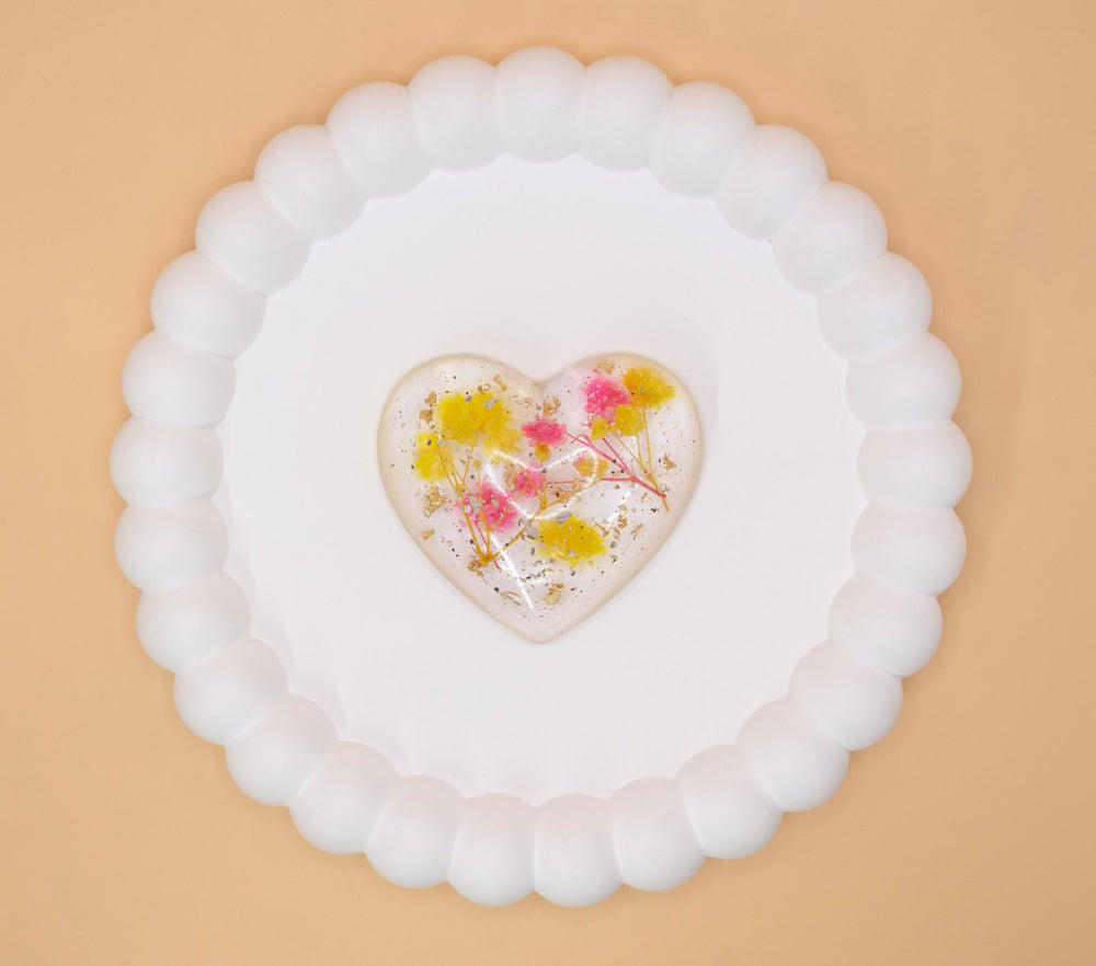 An overhead view of a heart shaped cremation ashes ornament, containing yellow and pink dried flowers and gold flakes, presented on a display tray