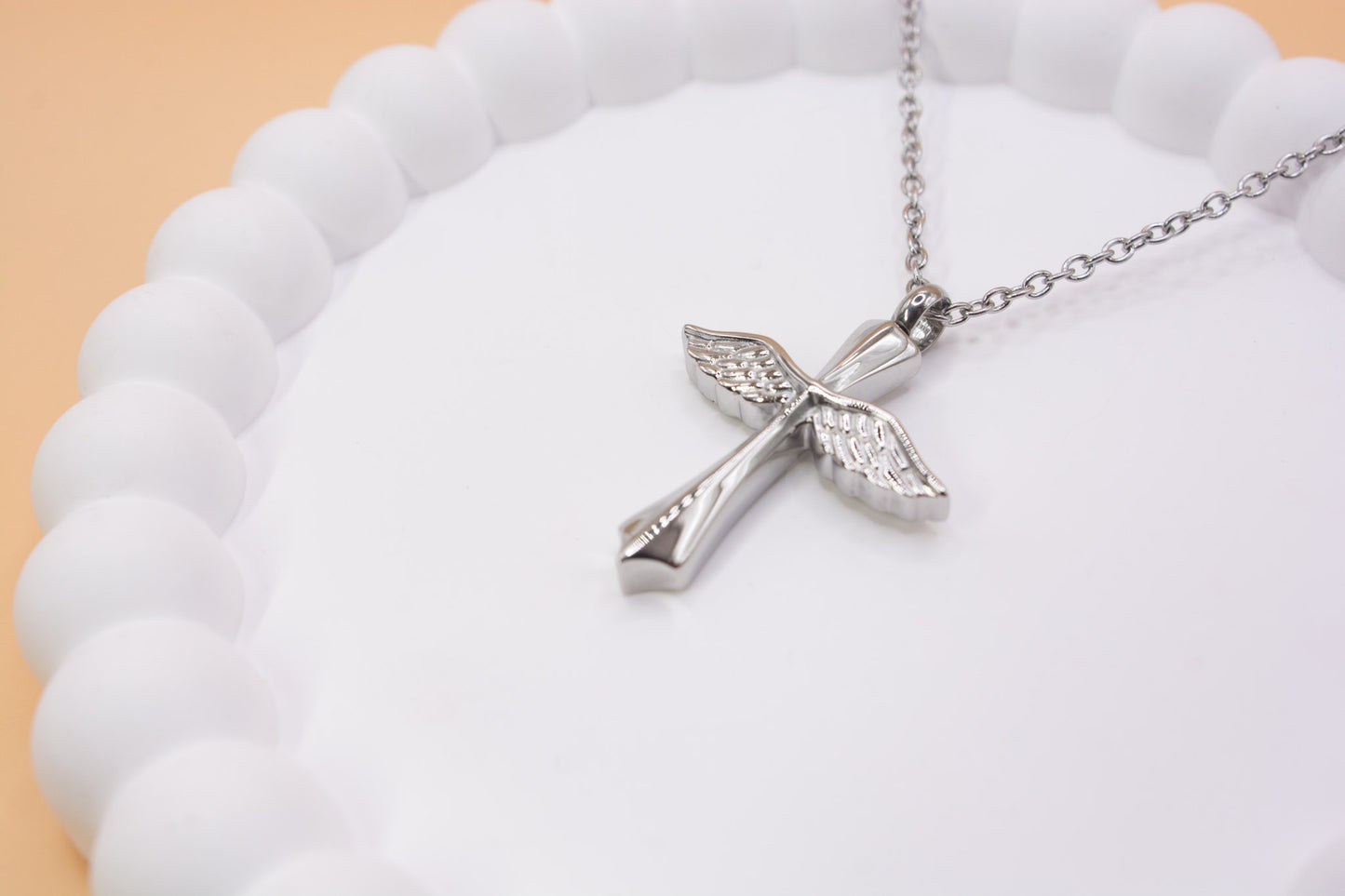 An angled close up view of a stainless steel cross with angel wings pendant, which can be self-filled with cremation ashes or sand from a favourite beach