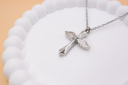 An angled close up view of a stainless steel cross with angel wings pendant, which can be self-filled with cremation ashes or sand from a favourite beach