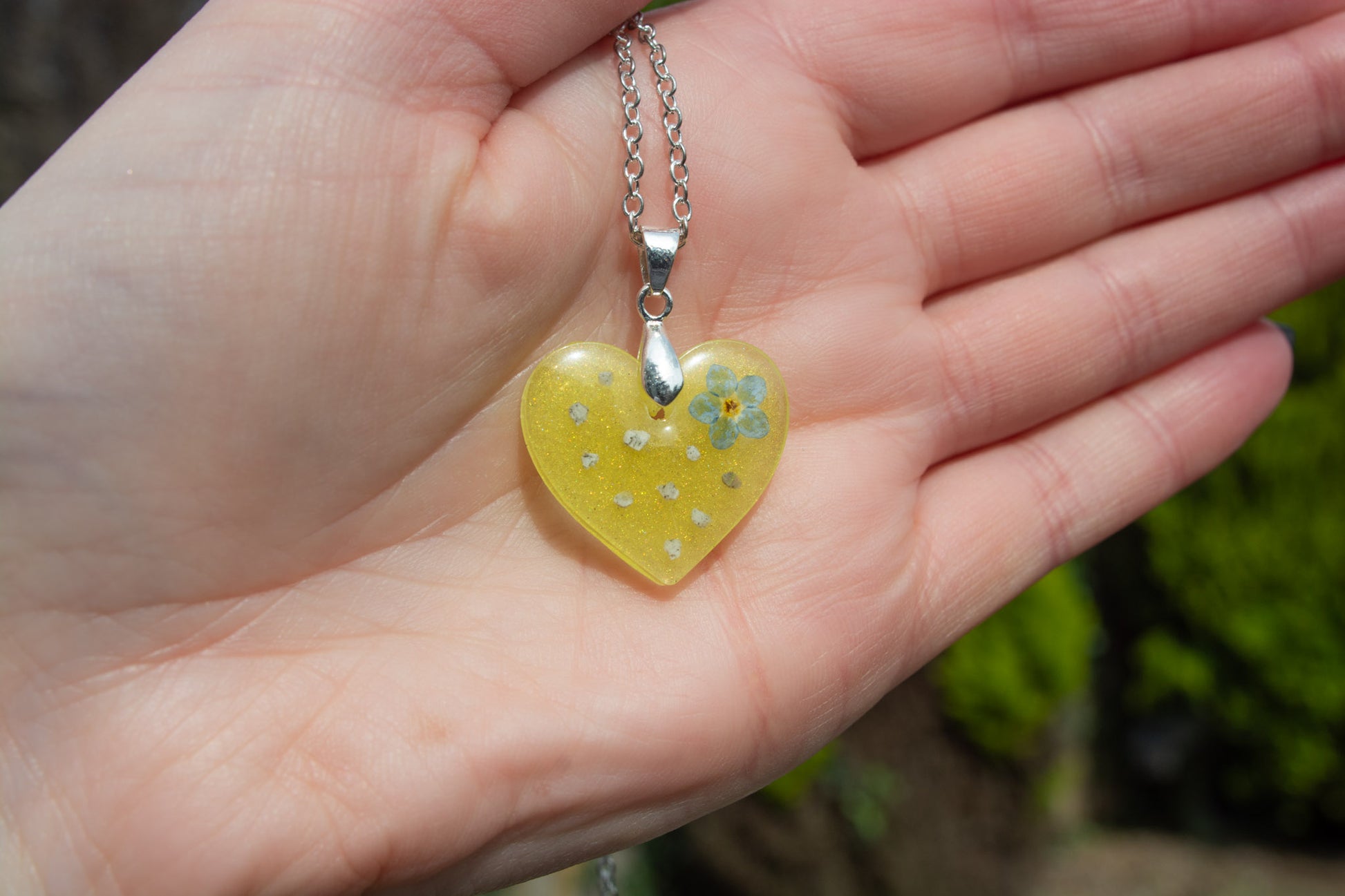 A hand showing a yellow heart keepsake memorial pendant outside, containing a Forget me Not flower and cremation ashes, set on a 925 sterling silver chain