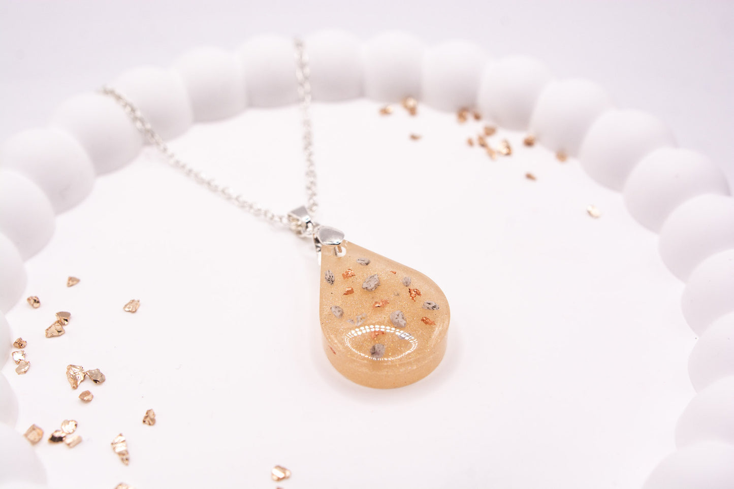 A close up, angled view of a metallic cream teardrop shaped keepsake memorial pendant containing cremation ashes and rose gold flakes, set on a 925 sterling silver chain