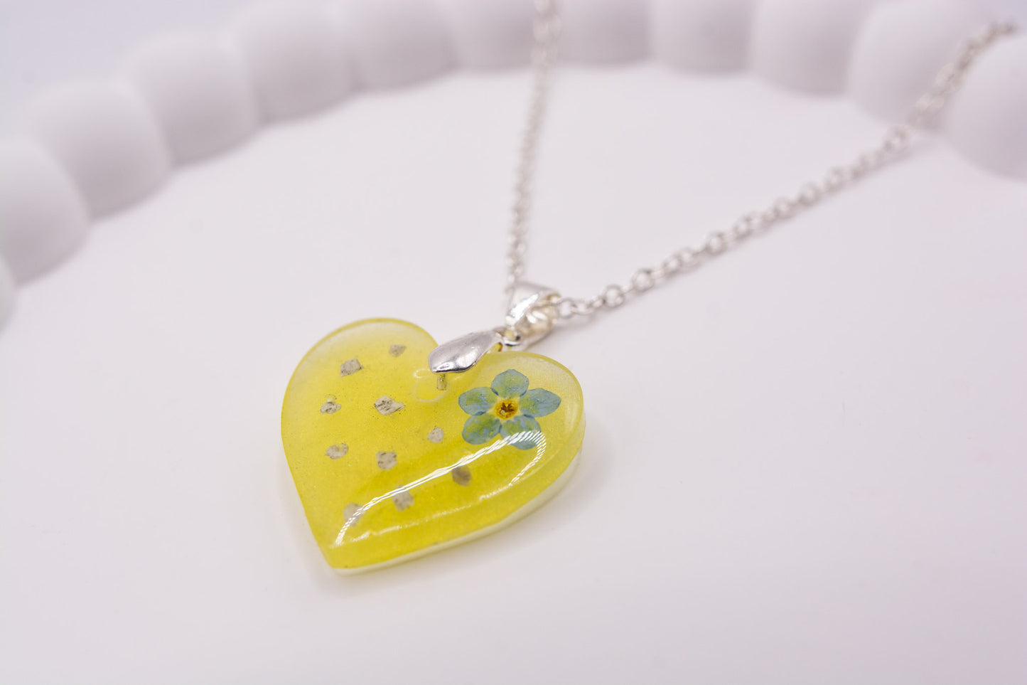 A close up angled view of a yellow heart keepsake memorial pendant, containing a real Forget me Not flower and cremation ashes, set on a 925 sterling silver chain