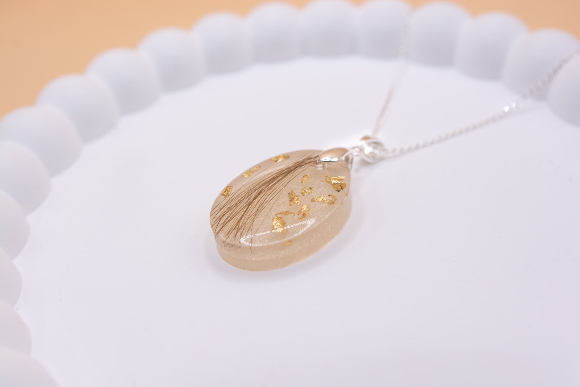 Angled close up view of an oval shaped memorial pendant, which contains a lock of hair and gold flakes, set on a 925 silver necklace chain, presented on a jewelry display tray