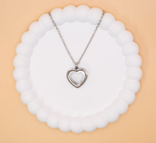 An aerial over head view of a stainless steel heart pendant, which can be self-filled with cremation ashes or sand from a favourite beach, it has a clear central window to see the contents