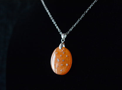 A dark background shot, showing an Oval shaped orange keepsake memorial pendant, containing cremation ashes and set on a 925 sterling silver chain