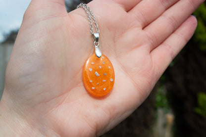 A hand presenting an oval shaped orange keepsake memorial pendant outside, containing cremation ashes and set on a 925 sterling silver chain