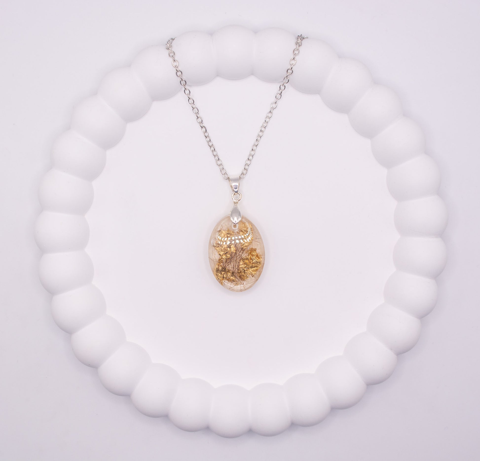 Over head view of a clear oval shaped keepsake memorial pendant, containing gold flake and human hair, set on a 925 sterling silver chain