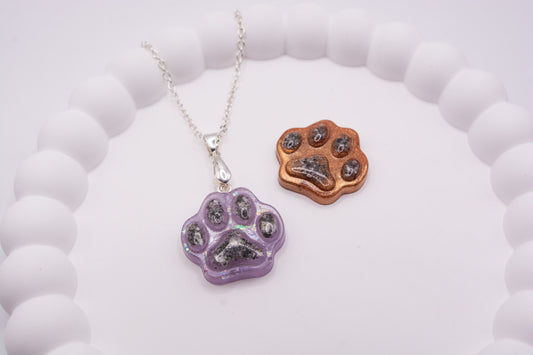 A close up angled view of 2 Paw print pendants containing pet cremation ashes, one is purple glitter and one is metallic brown, the purple one is presented on a 925 sterling silver chain