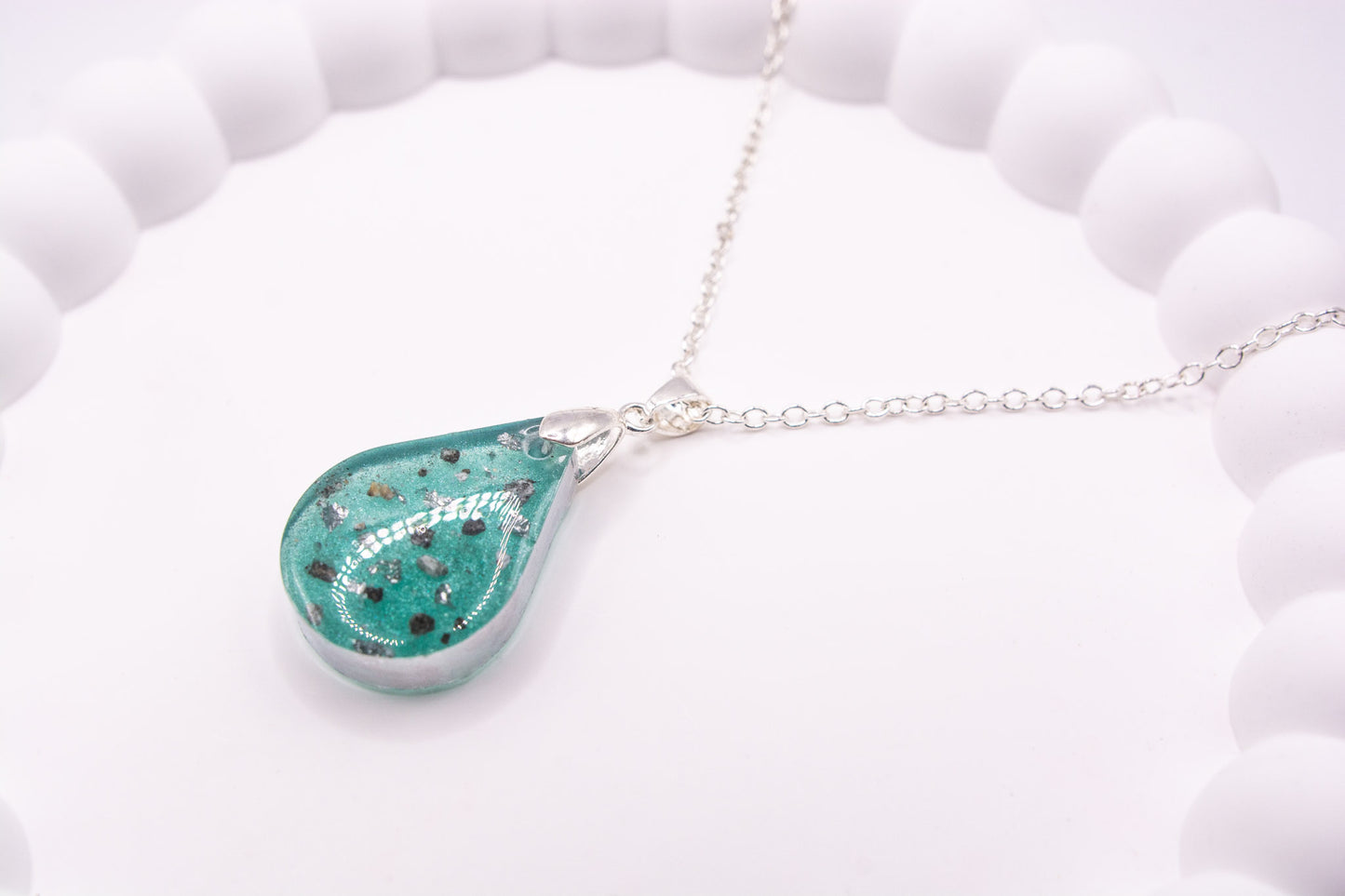 A closer up angled view of a teal teardrop shaped keepsake memorial pendant containing cremation ashes and silver flakes, set on a 925 sterling silver chain