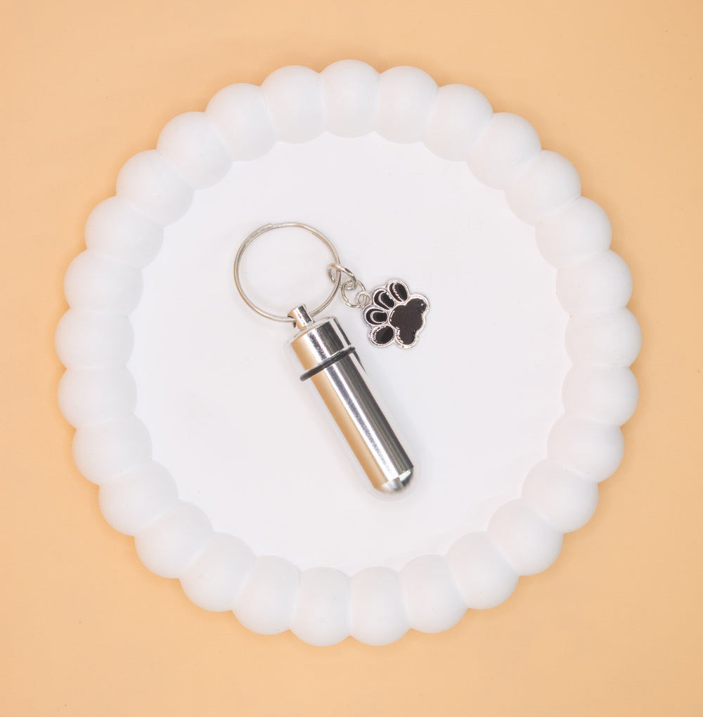 An aerial overhead view of a silver, stainless steel pet cremation ashes keyring, which can be self-filled with a lost loved cat dog rabbit etc cremation ashes ashes. Presented on a display tray.