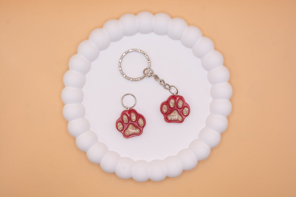 A view from above of 2 paw print shaped keepsakes. One is presented on a keying and the other is a dog collar tag. Both containing a lost loved pets cremation ashes.