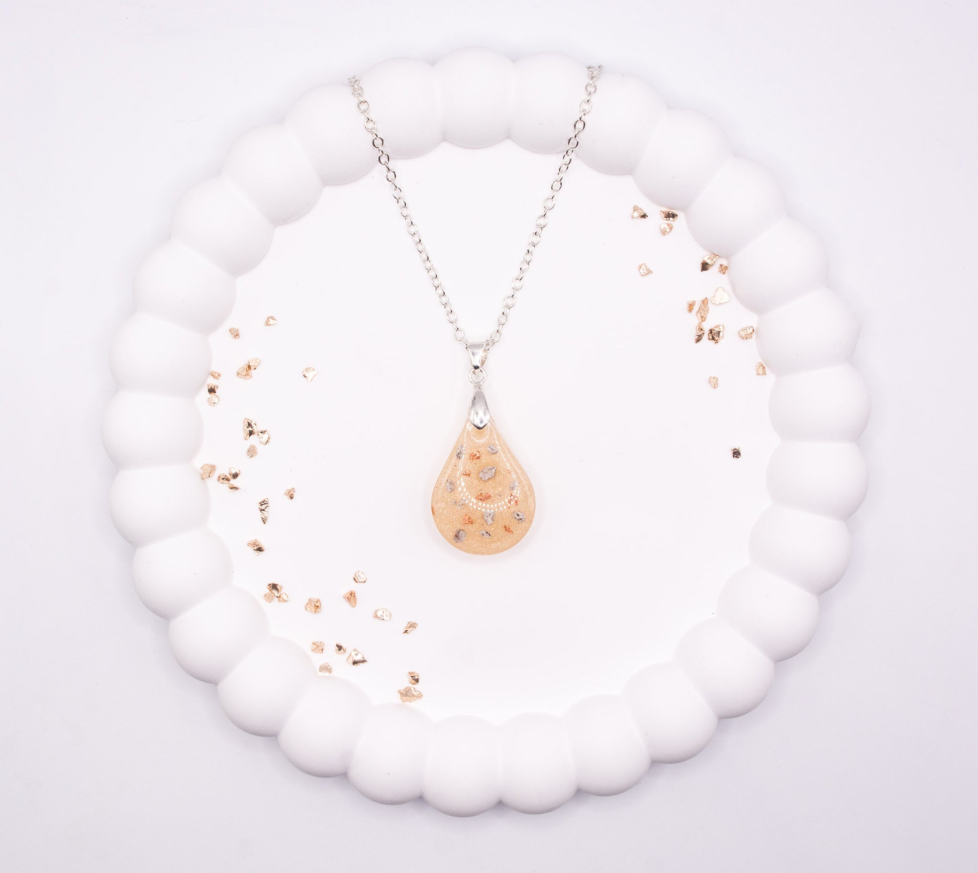 An aerial, over head view of a metallic cream teardrop shaped keepsake memorial pendant containing cremation ashes and rose gold flake, set on a 925 sterling silver chain and displayed on a jewelry tray