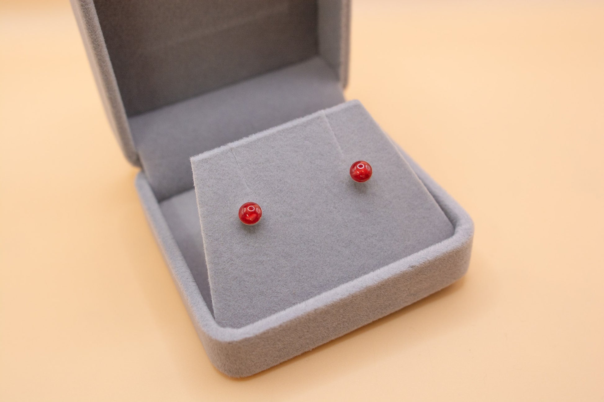 A close-up angled view of a plush grey jewelry box, inside are presented a pair of 925 sterling silver stud earrings with red resin and cremation ashes