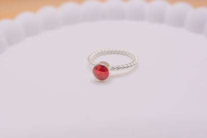 A close up shot of the 925 silver twist band ring, with a round bezel containing red resin and pet cremation ashes, on a white display tray