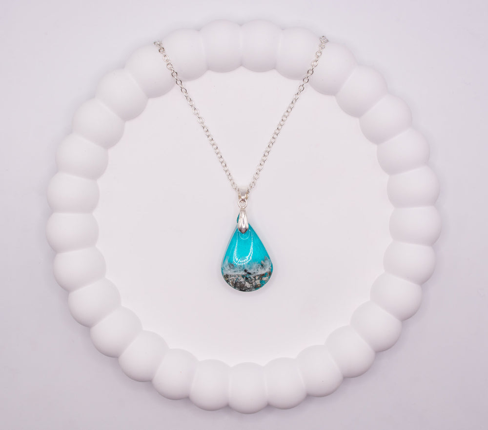 An aerial overhead view of a Teardrop shaped seashore memorial keepsake pendant, in sea blue, containing cremation ashes and beach sand, set on a sterling 925 silver chain and presented on a jewelry display tray